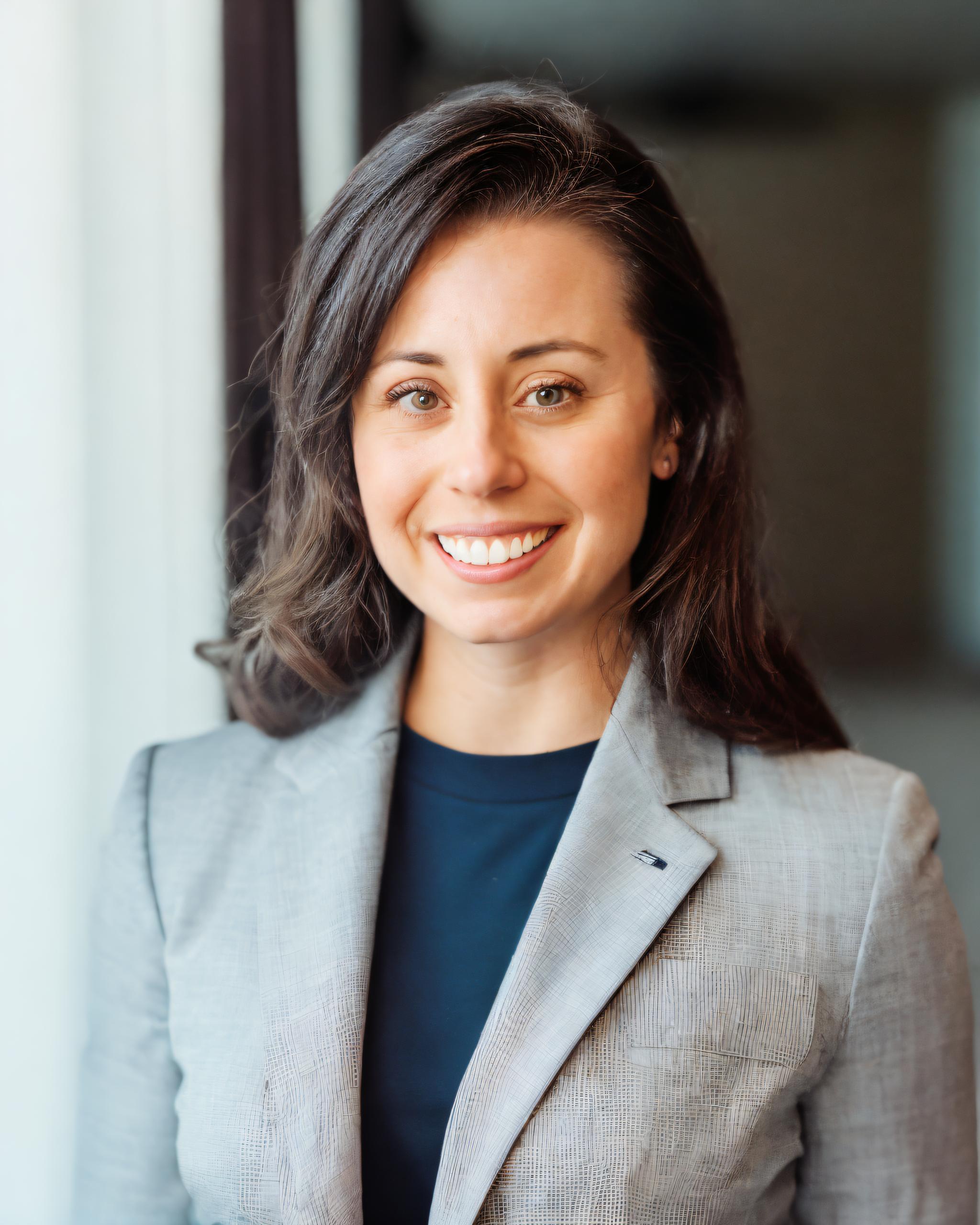 A smiling woman in a professional gray blazer and navy shirt stands against a blurred background