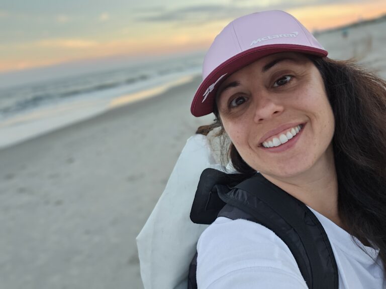 Smiling woman wearing a pink McLaren cap stands on a beach at sunset